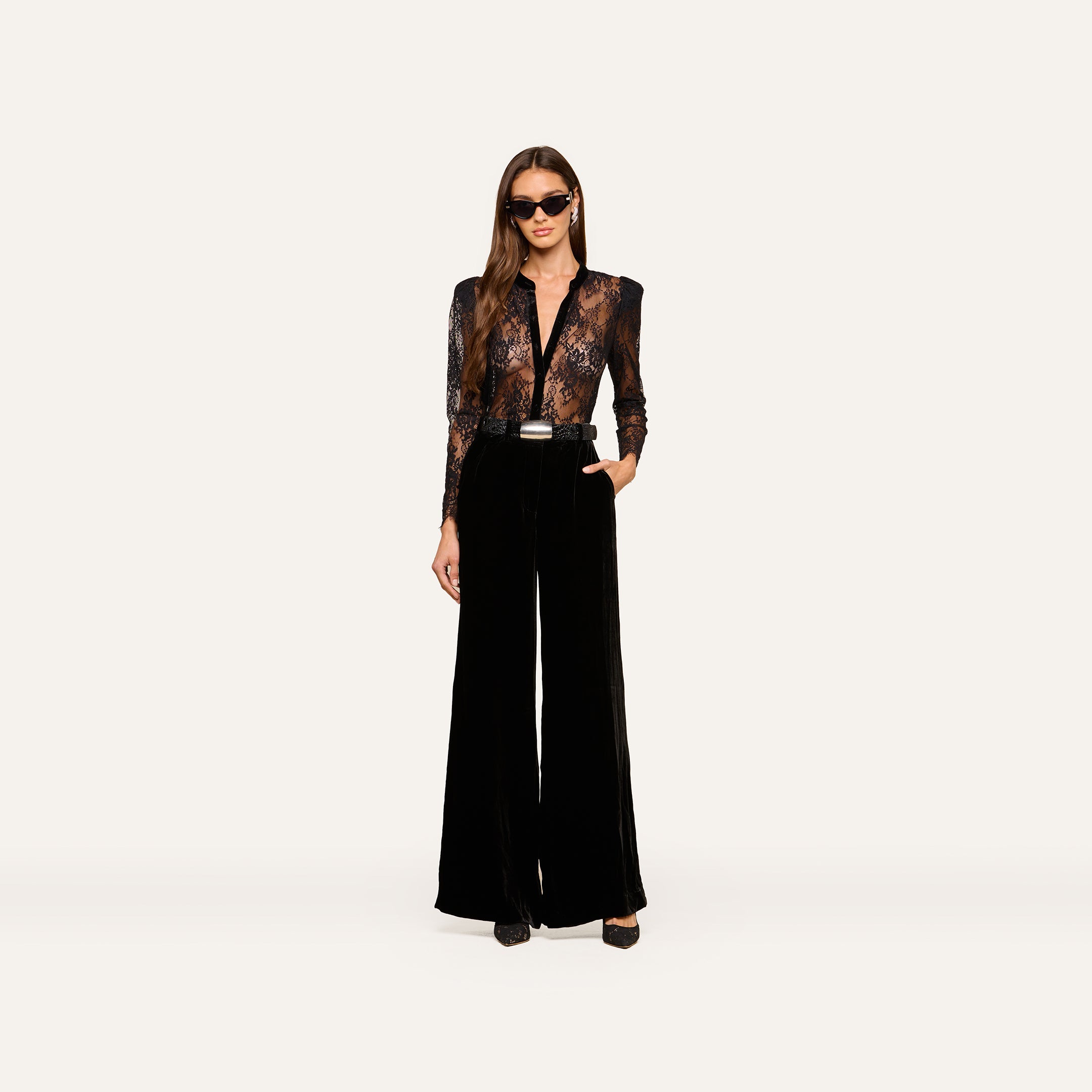 Wearing the Dorothea Velvet Lace Blouse, a woman in sunglasses stands confidently with one hand in her pocket, showcasing its sheer Chantilly lace long sleeves tucked into high-waisted, wide-leg black velvet pants with elegant velvet seams and finished with black shoes.