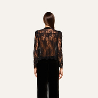 A woman with straight brown hair faces away, wearing the Dorothea Velvet Lace Blouse—crafted from sheer Chantilly lace with long sleeves—and black pants, set against a plain light background.