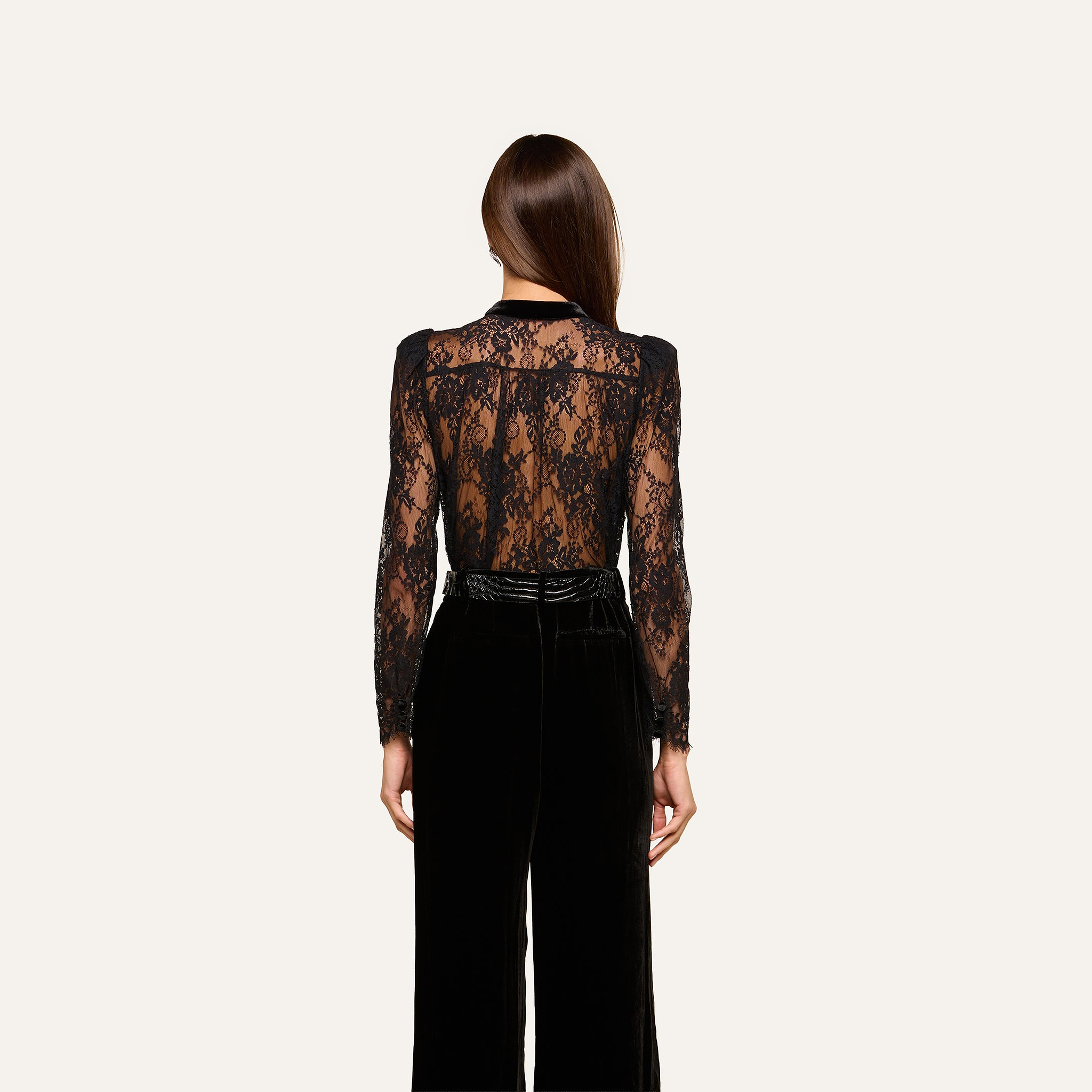 A woman facing away wears the Dorothea Velvet Lace Blouse—a sheer black Chantilly lace long-sleeve top—paired with high-waisted black velvet pants accented by delicate velvet seams. Her straight brown hair is down, and the background is plain off-white.
