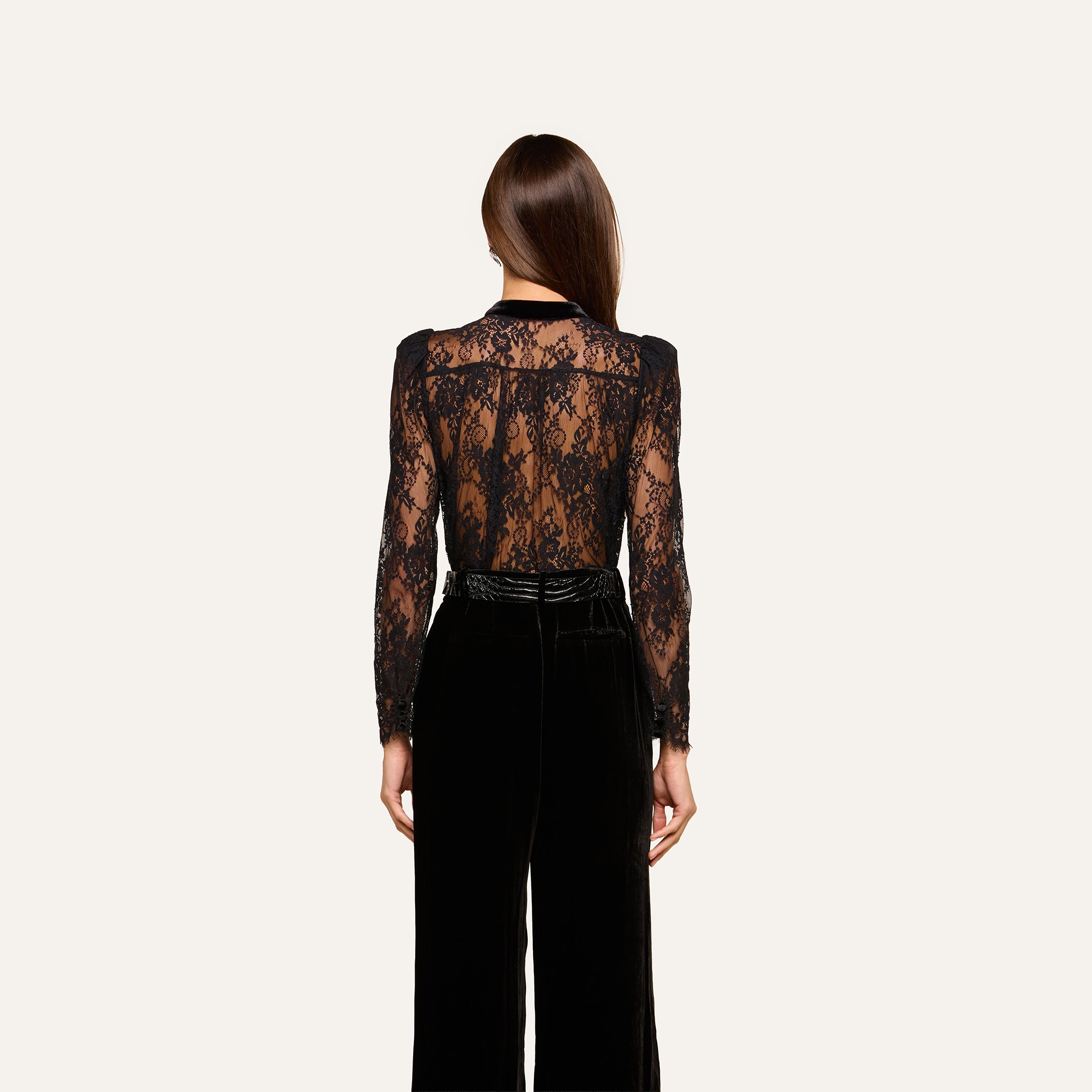 A woman facing away wears the Dorothea Velvet Lace Blouse—a sheer black Chantilly lace long-sleeve top—paired with high-waisted black velvet pants accented by delicate velvet seams. Her straight brown hair is down, and the background is plain off-white.