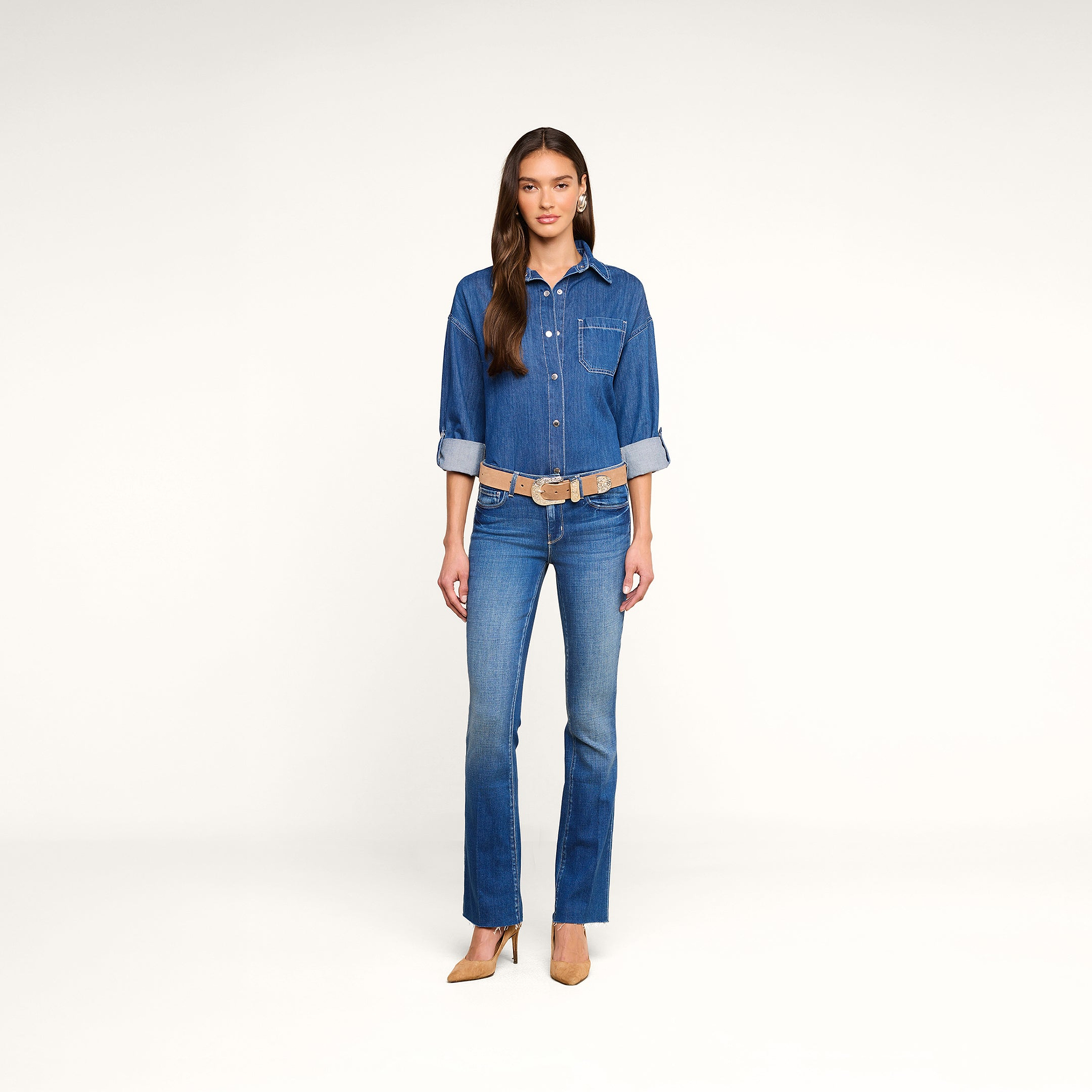 A woman stands facing forward, wearing the Jorah Oversized Shirt in featherweight indigo denim paired with blue jeans and tan high heels. The background is plain white.