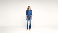 A woman stands against a plain white background, wearing a denim shirt with rolled-up sleeves, blue jeans, tan heels, and a tan belt, facing forward with a neutral expression.