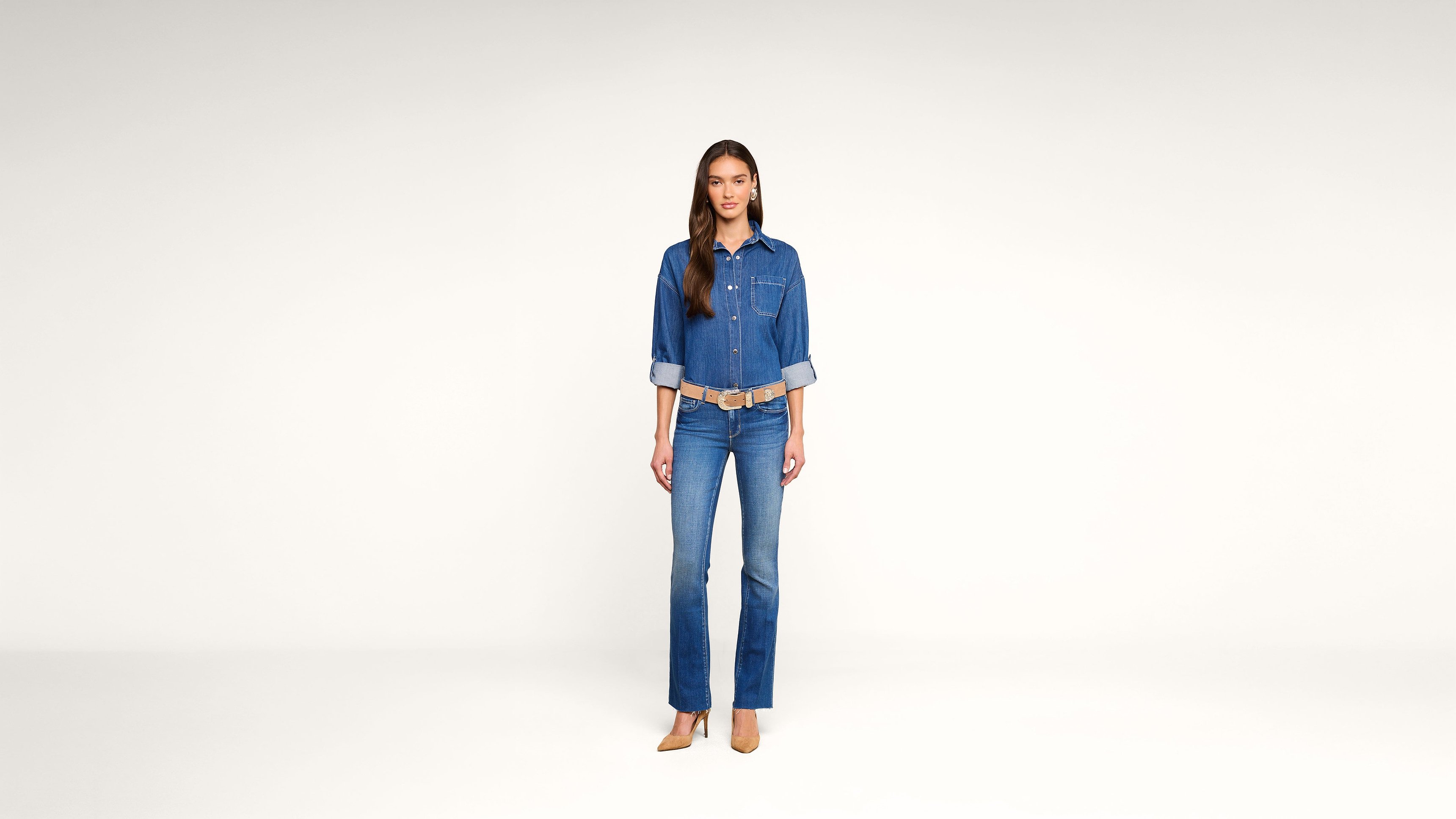 A woman stands against a plain white background, wearing a denim shirt with rolled-up sleeves, blue jeans, tan heels, and a tan belt, facing forward with a neutral expression.