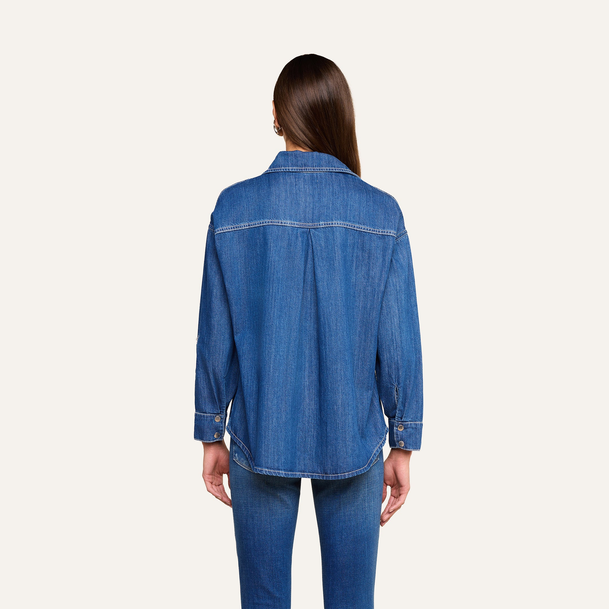 A person with long brown hair stands facing away, wearing the Jorah Oversized Shirt in indigo featherweight denim and matching blue jeans against a plain light background.