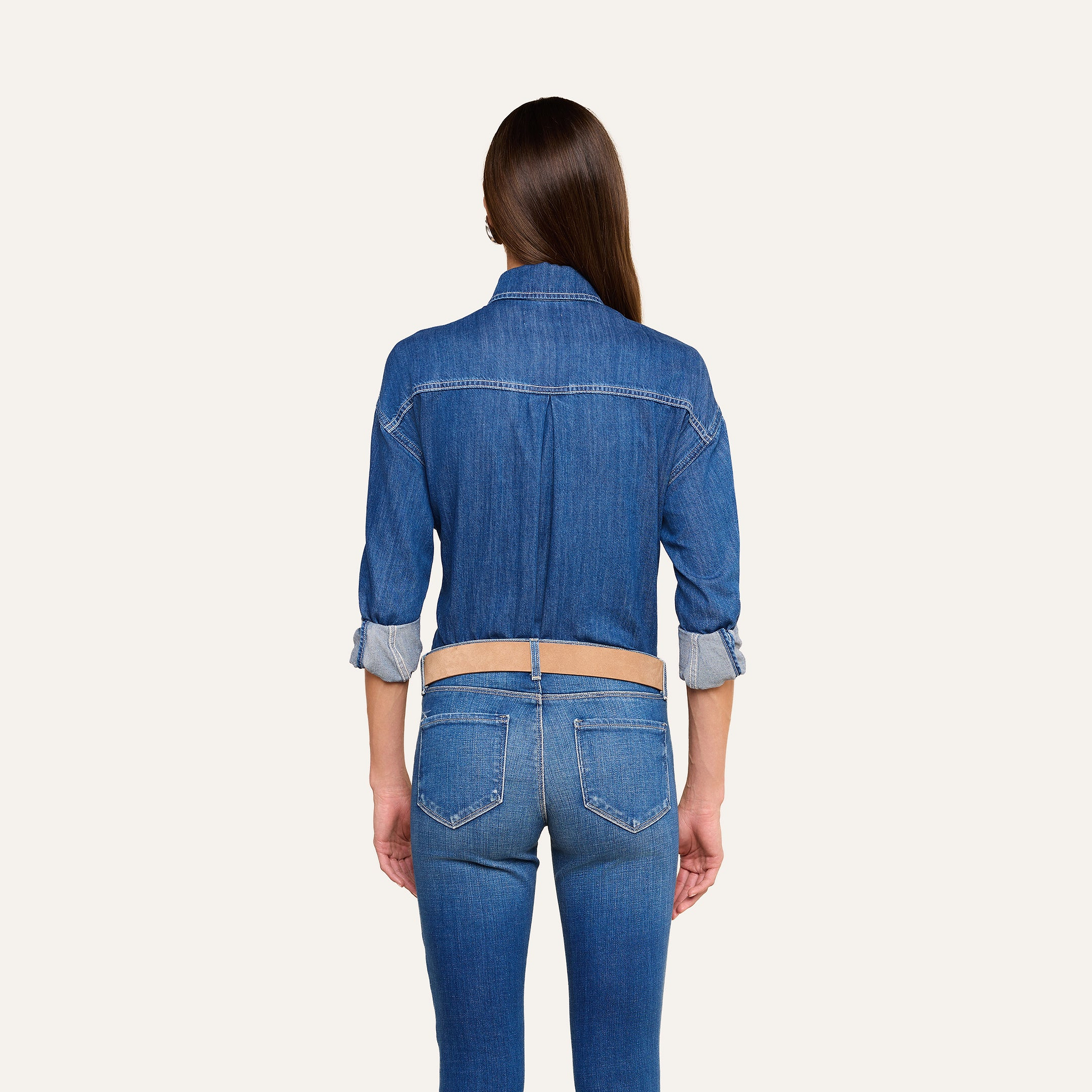 A woman with long brown hair stands facing away, wearing the Jorah Oversized Shirt with rolled-up sleeves and matching indigo blue jeans against a plain light background.