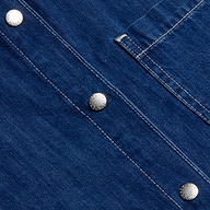 Close-up of the indigo featherweight denim fabric on the Jorah Oversized Shirt, featuring visible white stitching, three silver snap buttons, and part of its oversized pocket flap.