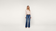 A woman stands against a plain white background, wearing a light pink blouse tucked into high-waisted blue wide-leg jeans and open-toed heels. She has long, wavy blonde hair and a neutral expression.