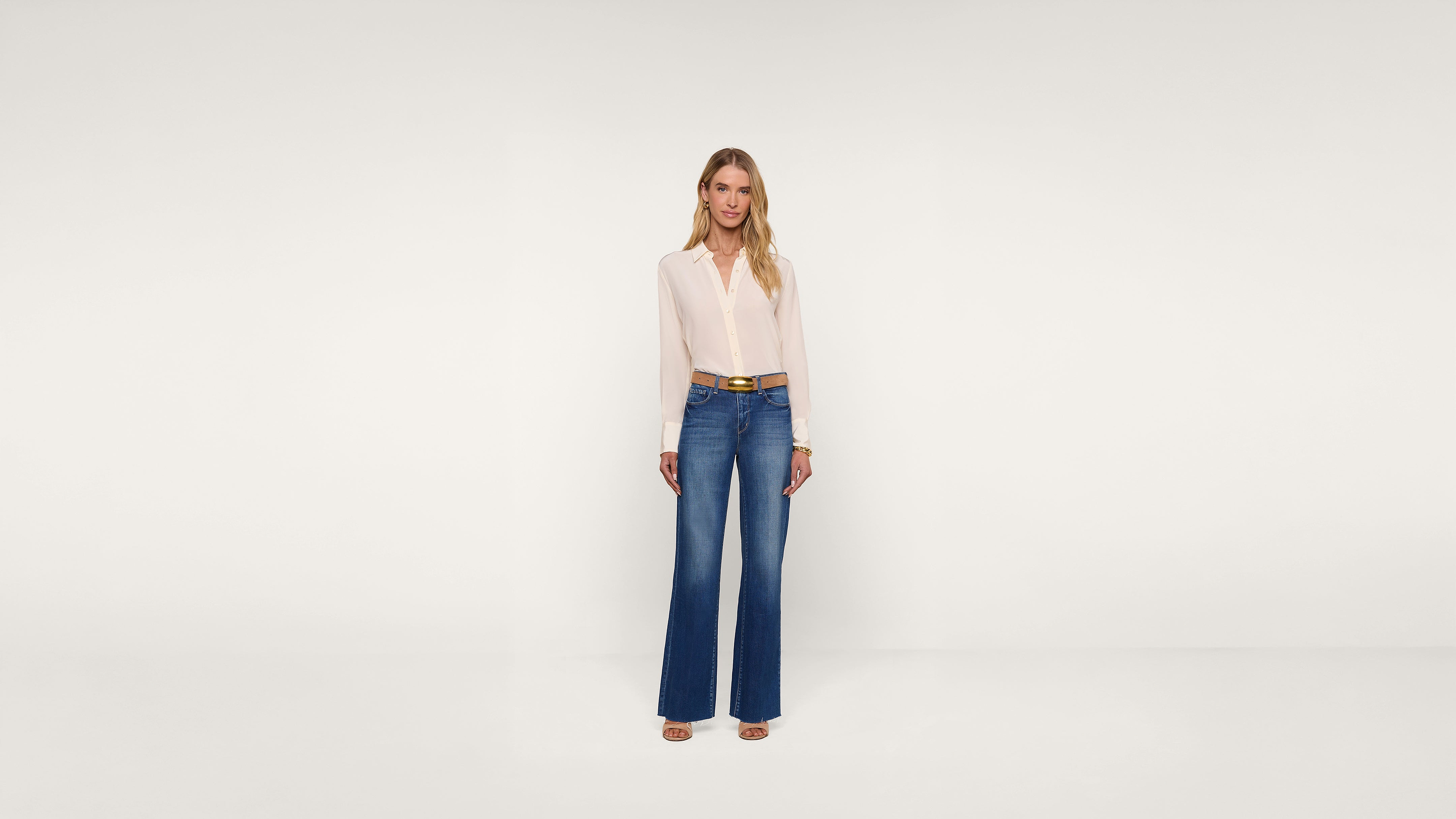 A woman stands against a plain white background, wearing a light pink blouse tucked into high-waisted blue wide-leg jeans and open-toed heels. She has long, wavy blonde hair and a neutral expression.
