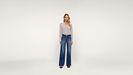 A woman stands facing forward against a plain white background, wearing a light-colored blouse, blue wide-leg jeans, black belt, and black sandals. She has long blonde hair and a neutral expression.
