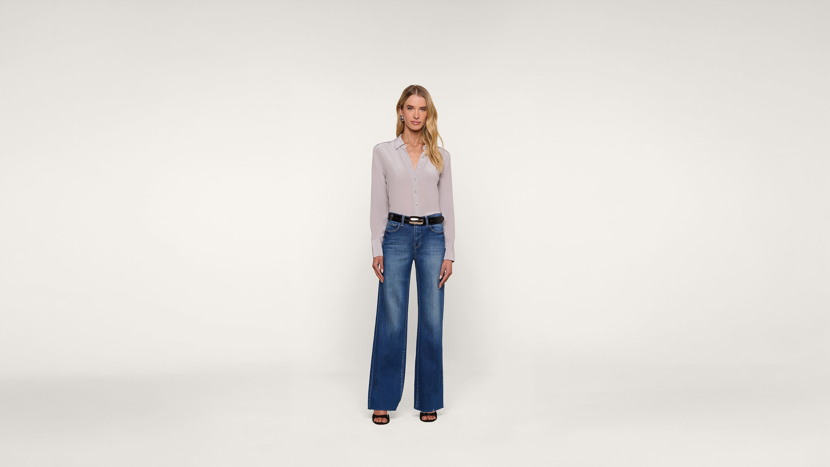 A woman stands facing forward against a plain white background, wearing a light-colored blouse, blue wide-leg jeans, black belt, and black sandals. She has long blonde hair and a neutral expression.