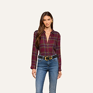 A woman with long brown hair wears the Suzette Shirt—a maroon plaid button-down with a high-low hemline—tucked into blue jeans with a black belt, standing against a plain light background.