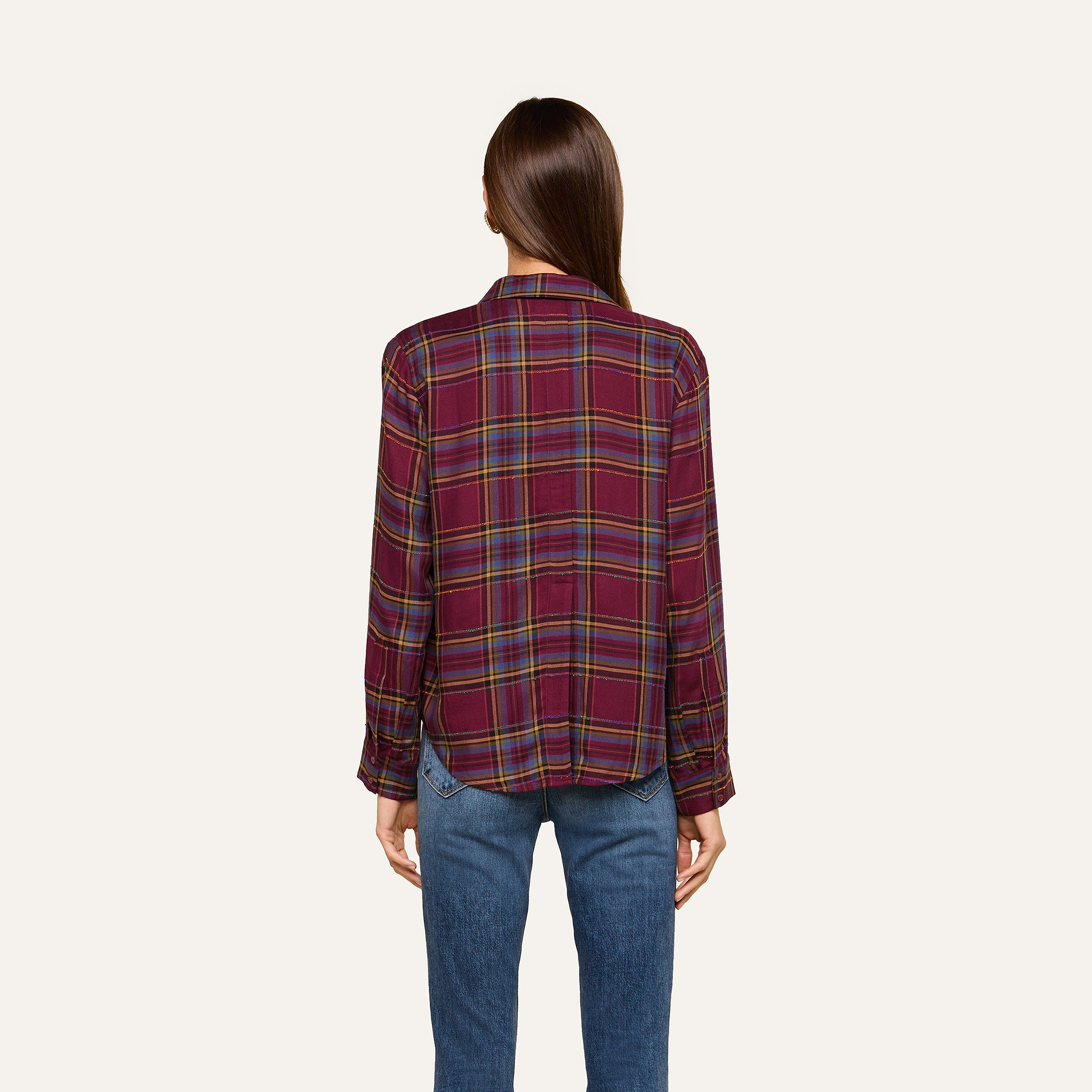 A woman stands with her back to the camera against a plain light background, wearing the Suzette Shirt—a red plaid button-down with a high-low hemline—paired with blue jeans.