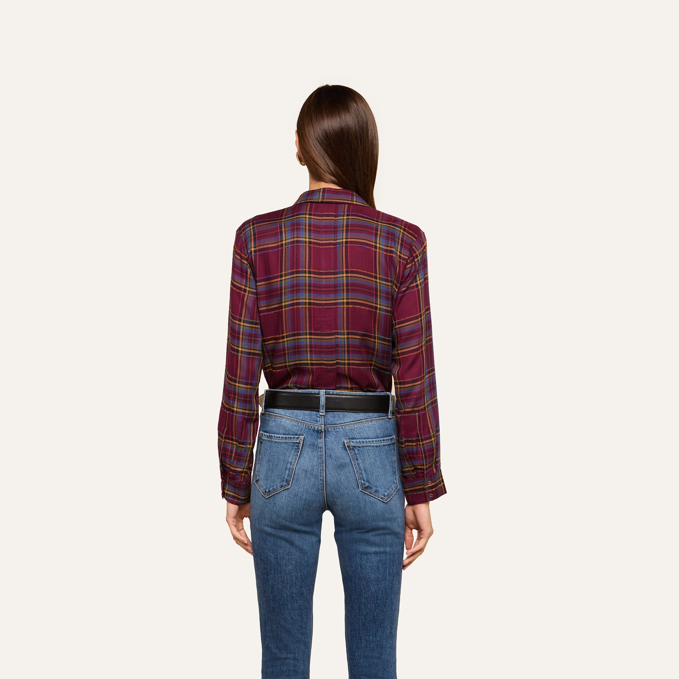 A woman with straight brown hair is standing facing away, wearing the Suzette Shirt tucked into high-waisted blue jeans. The background is plain and light-colored.