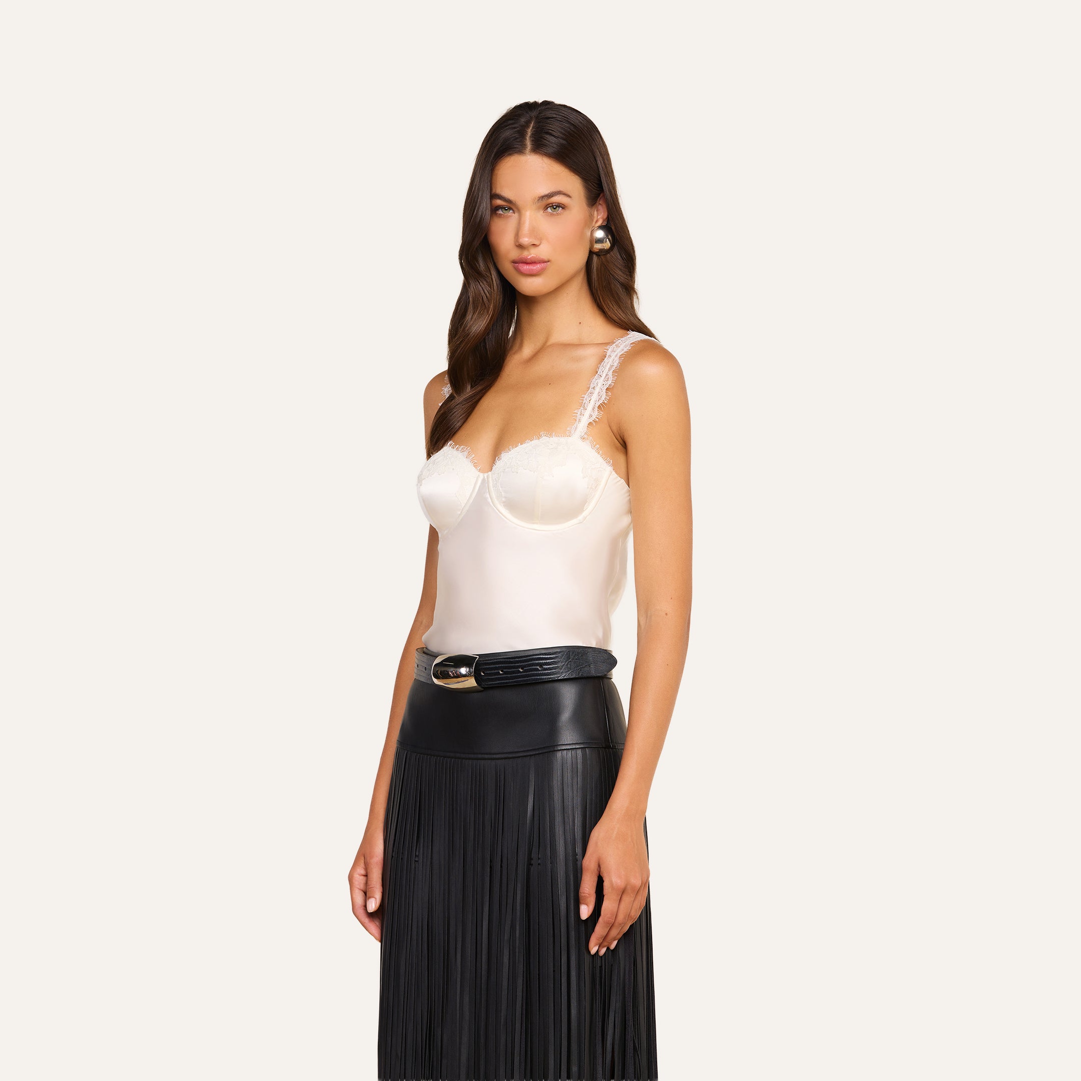 Wearing the Soleia Lace Camisole, a woman stands facing forward against a plain light background, paired with a black pleated leather skirt.