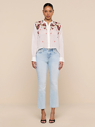 A woman stands facing forward against a plain white background wearing the Philomena Blouse, a semi-sheer white Ramie top with floral embroidery, paired with light blue jeans and black heeled sandals.