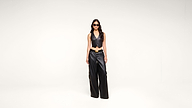 A woman wearing sunglasses, a black leather vest, and matching wide-leg pants stands confidently against a plain white background.