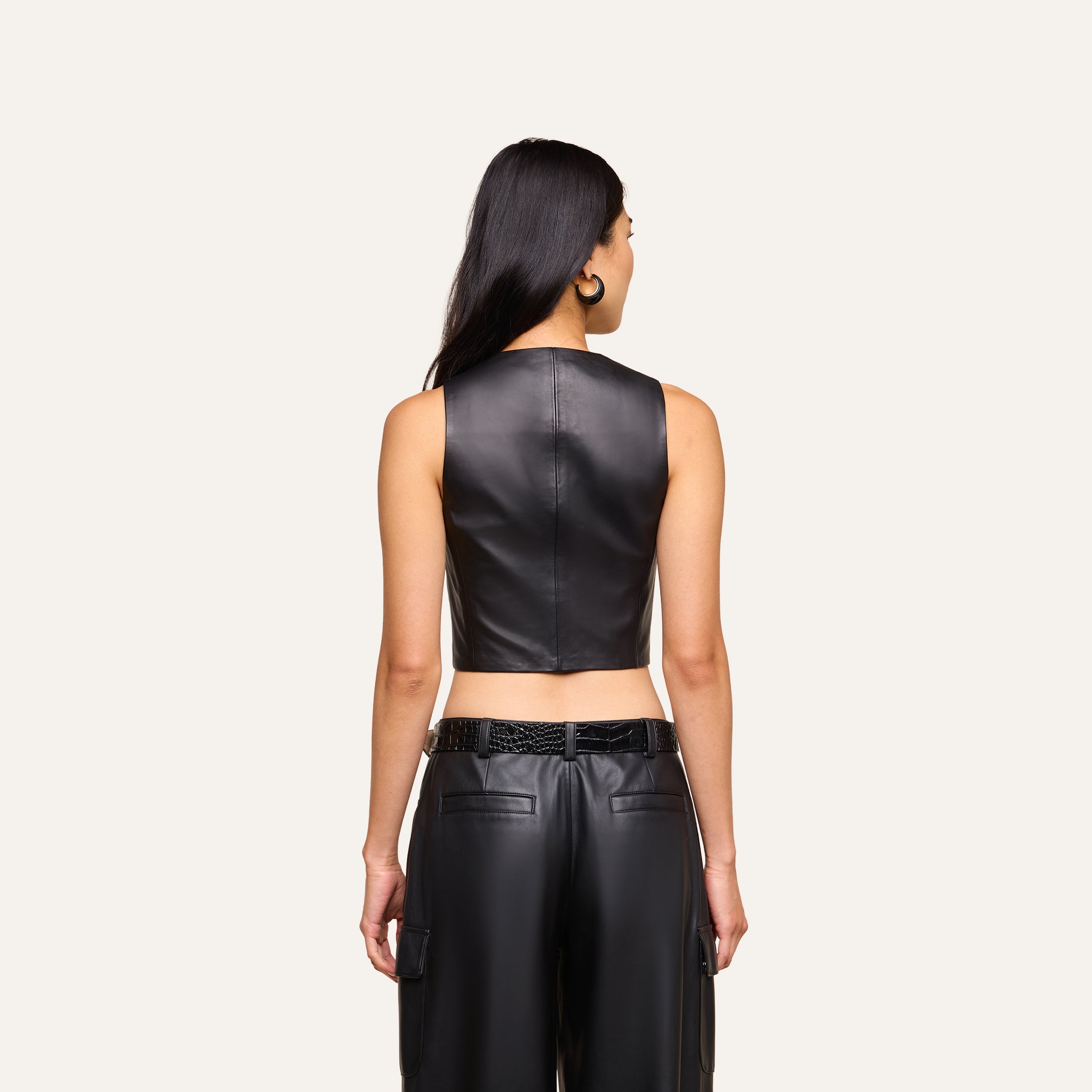 A woman with long dark hair, seen from behind, wears the sleeveless black Jianna Leather Vest paired with black leather pants against a plain light background—an edgy look inspired by Fall 2026 fashion.