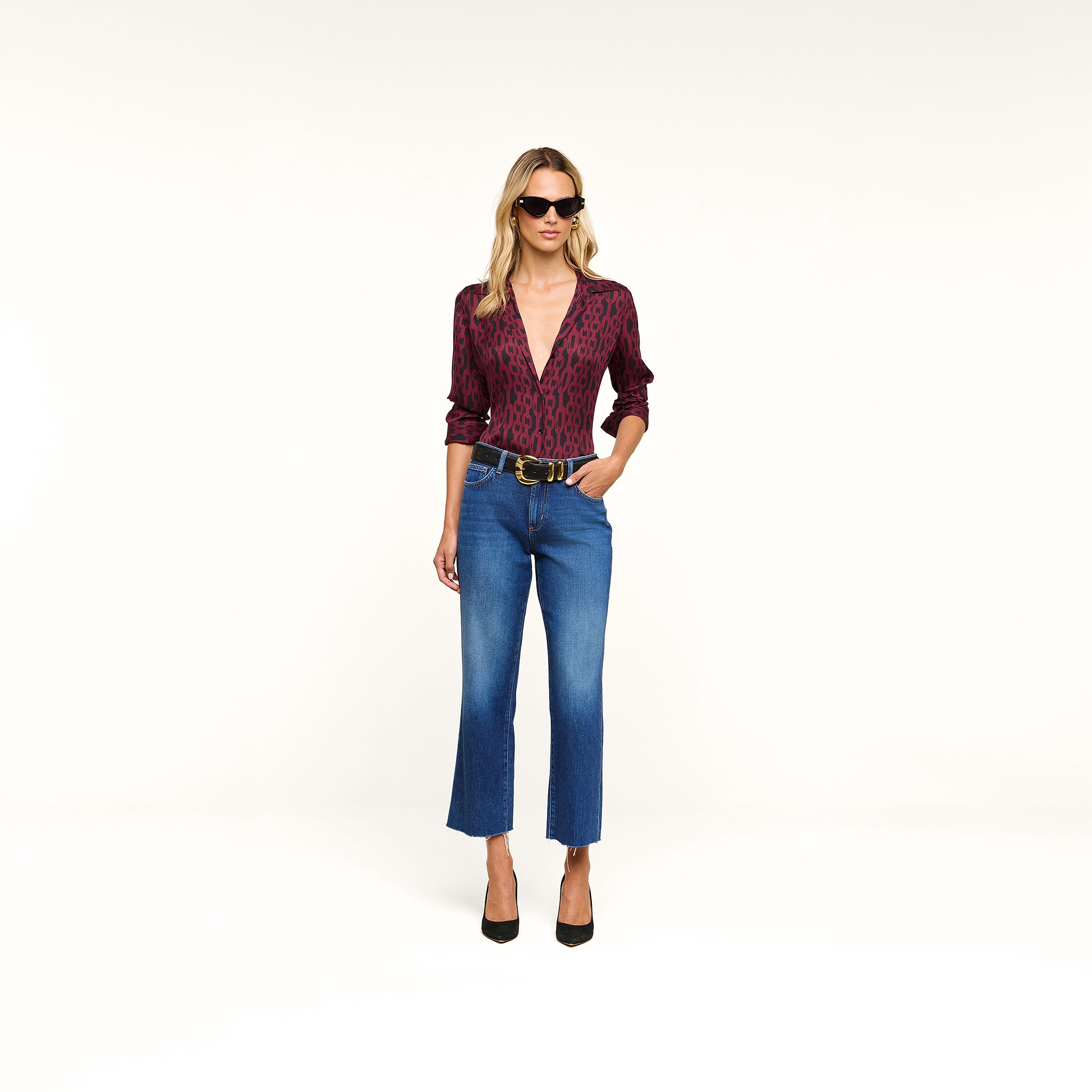 A woman wearing sunglasses and the Nina Silk Blouse—a deep V-neck maroon leopard print blouse with a relaxed fit—paired with blue cropped jeans, a black belt, and black heels stands against a plain white background.