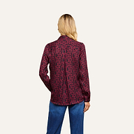 A person with long blonde hair is seen from behind against a plain light background, wearing the Nina Silk Blouse—a burgundy patterned button-down shirt—paired with relaxed-fit blue jeans.