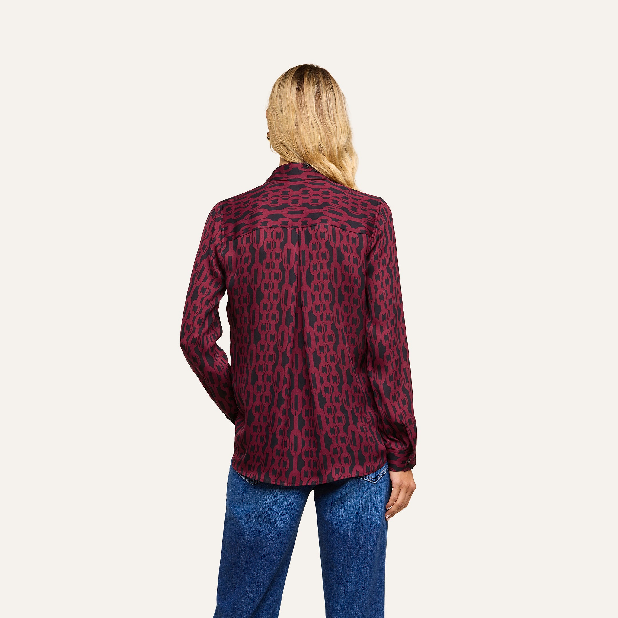 A person with long blonde hair is seen from behind against a plain light background, wearing the Nina Silk Blouse—a burgundy patterned button-down shirt—paired with relaxed-fit blue jeans.