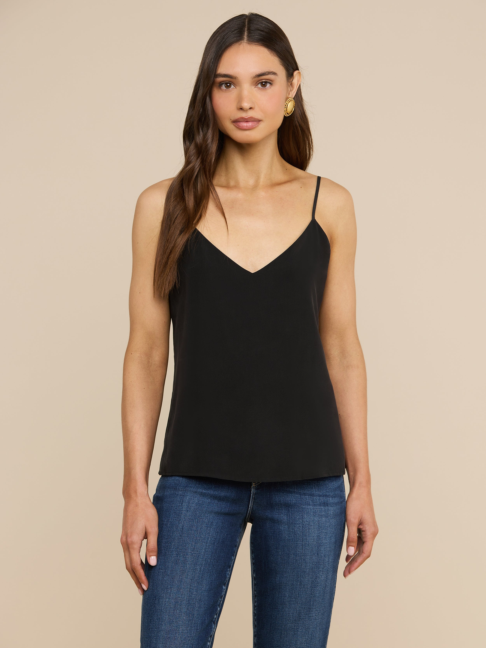 Una donna dai lunghi capelli castani indossa la canotta Jane Silk di colore nero, caratterizzata da spalline regolabili, abbinata a un paio di jeans blu. È in piedi, rivolta in avanti, su uno sfondo beige a tinta unita, con un’espressione neutra.