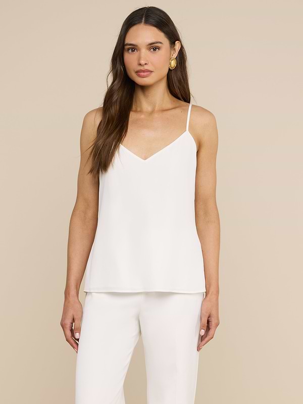 Una donna dai lunghi capelli castani indossa la canotta Jane Silk, un top bianco in seta dal taglio al diagona con spalline regolabili, abbinato a pantaloni bianchi coordinati, mentre posa davanti a uno sfondo beige.
