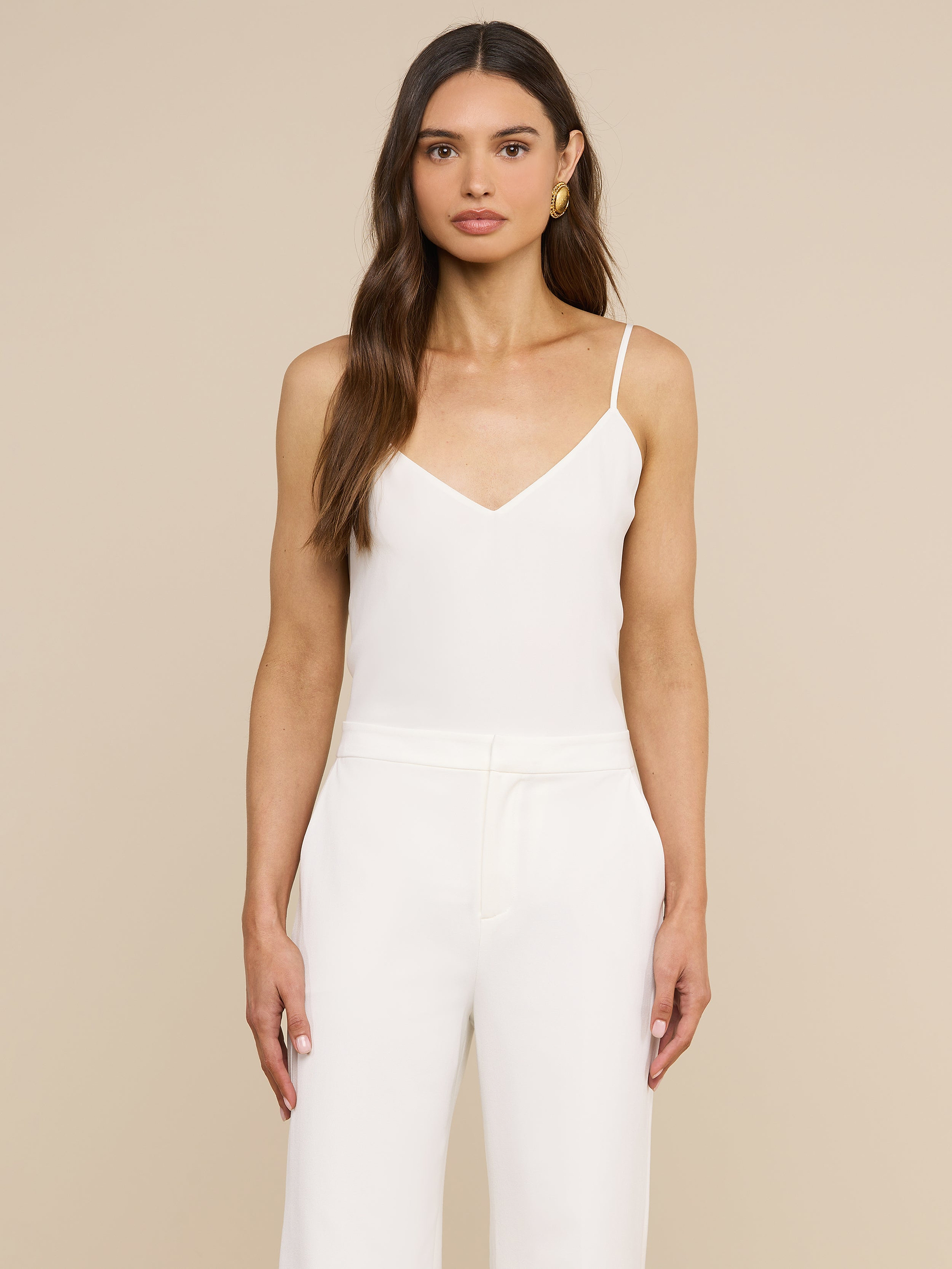 Eine Frau mit langen braunen Haaren und goldenen Creolen steht vor einem beigen Hintergrund und trägt das Jane Silk Camisole mit verstellbaren Trägern sowie eine dazu passende weiße Hose. Ihr Gesichtsausdruck ist neutral.