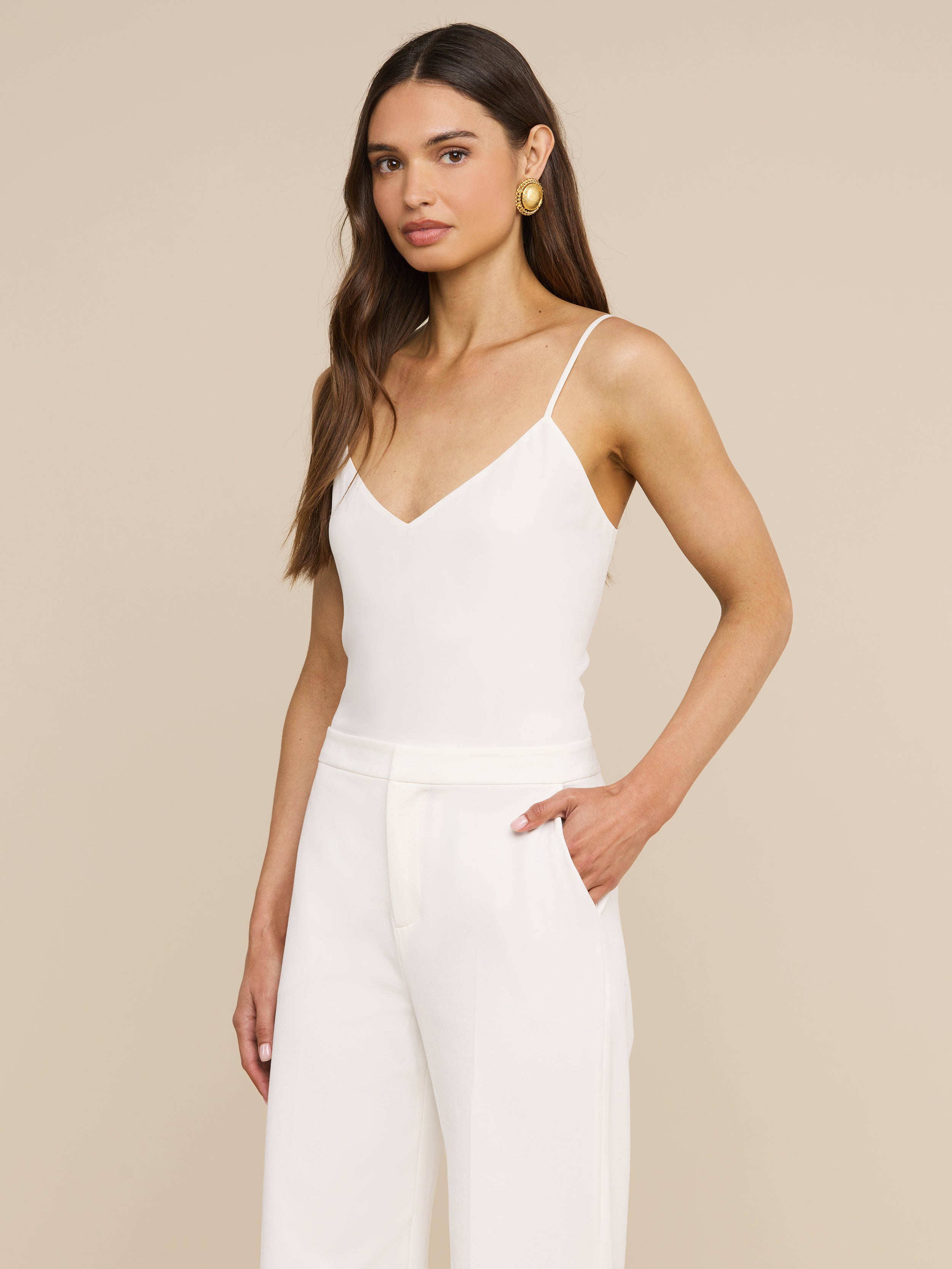 Una donna dai lunghi capelli castani e con orecchini doro indossa la canotta bianca Jane Silk con spalline regolabili e pantaloni a vita alta; in piedi su uno sfondo beige, con una mano in tasca, guarda verso la fotocamera.