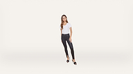 Woman standing against a plain white background, wearing a white t-shirt, black skinny jeans, and black high-heeled sandals. She has long brown hair and a neutral expression, with one hand at her side and the other on her thigh.