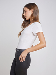 Wearing the Cory Cotton Scoopneck Tee, a woman stands in profile against a plain light background, her left hand tucked in the pocket of black pants and her long hair loose.