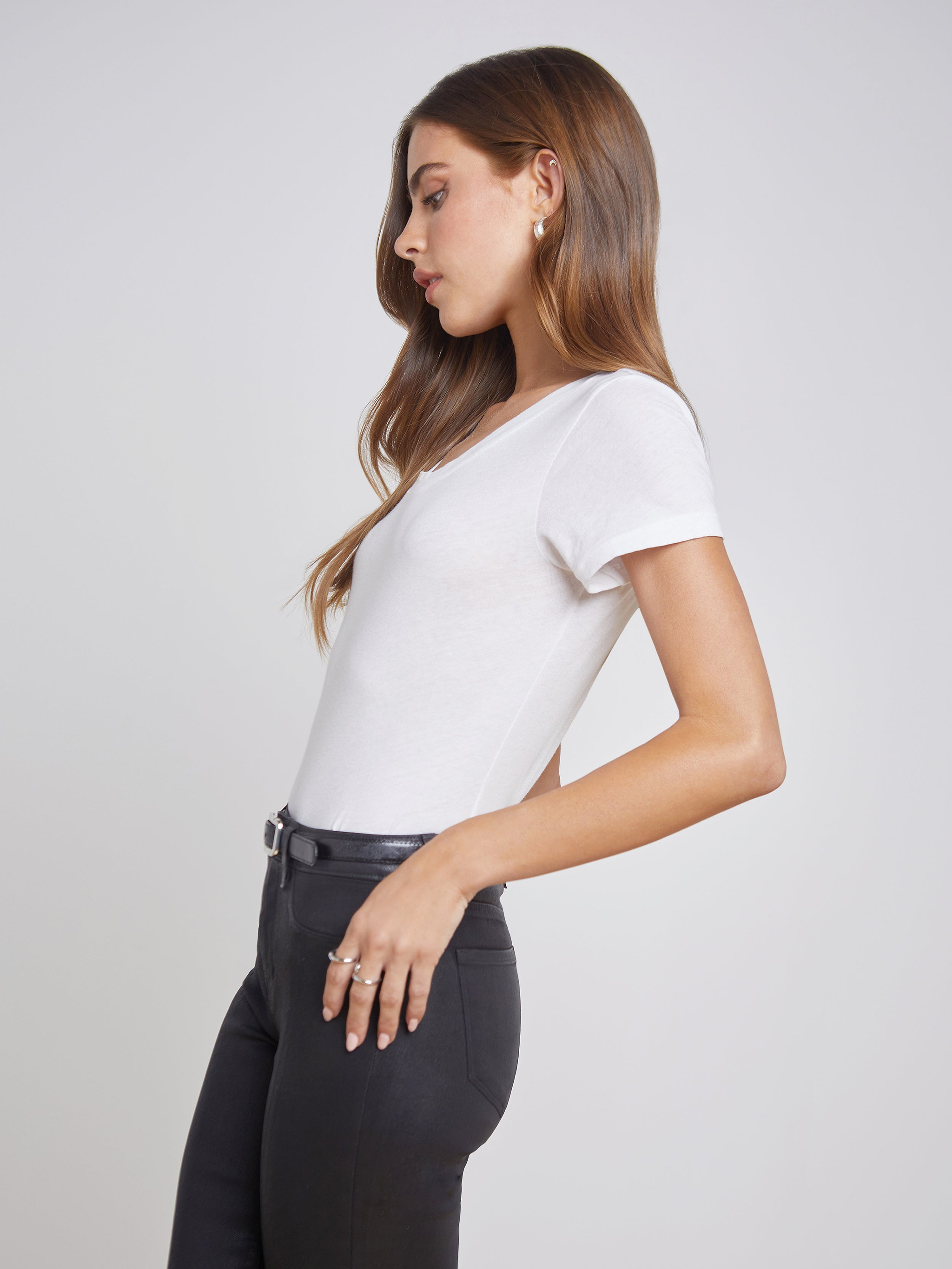 Wearing the Cory Cotton Scoopneck Tee, a woman stands in profile against a plain light background, her left hand tucked in the pocket of black pants and her long hair loose.