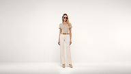 A woman wearing sunglasses, a beige short-sleeve top, wide-leg pants, a tan belt, and matching shoes stands against a plain white background.