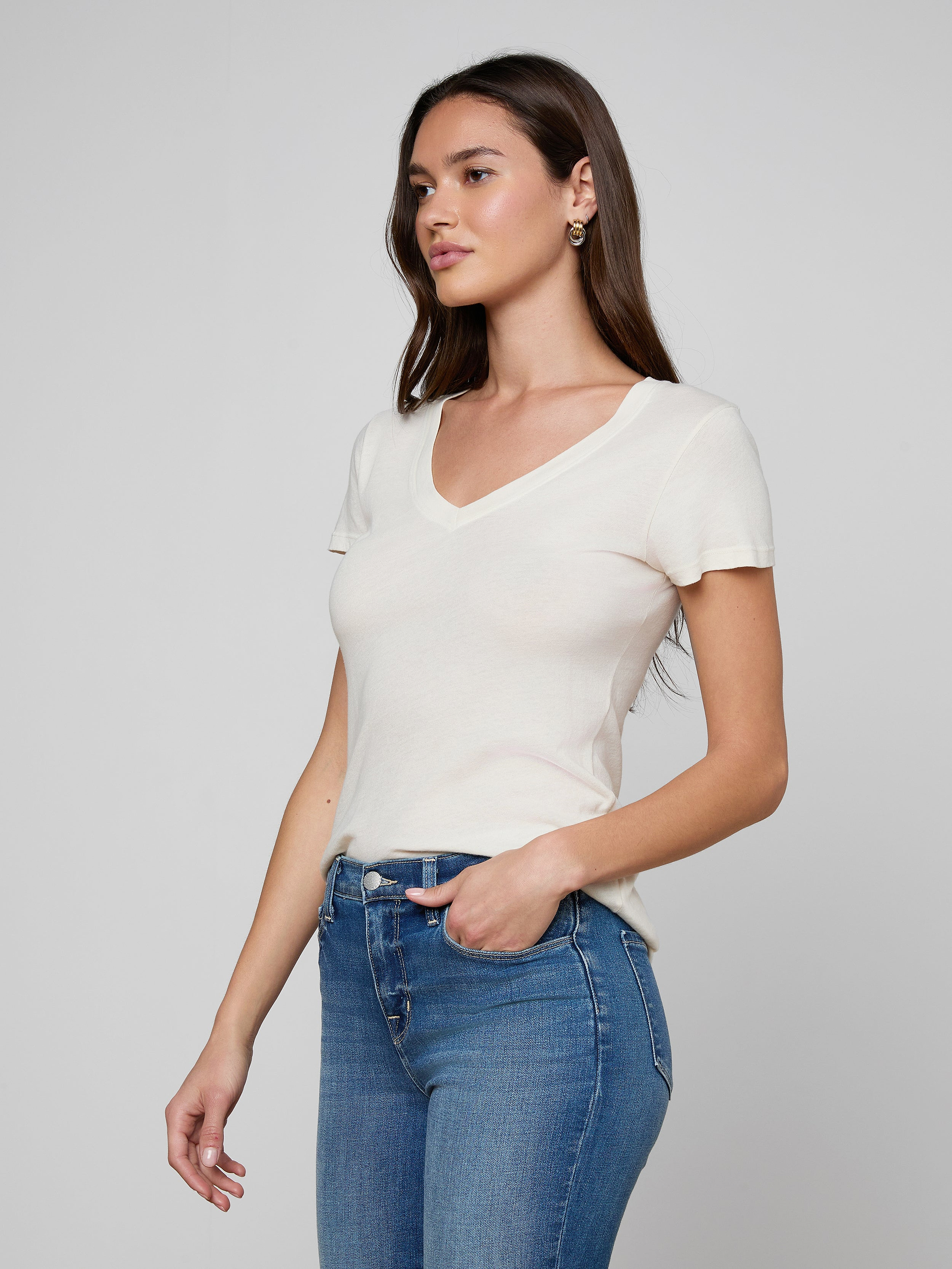 A woman with long blonde hair wears the Becca Cotton V-Neck Tee in a vintage off-white shade, paired with black pants, a black belt featuring a gold buckle, and large round earrings, standing against a plain light background.