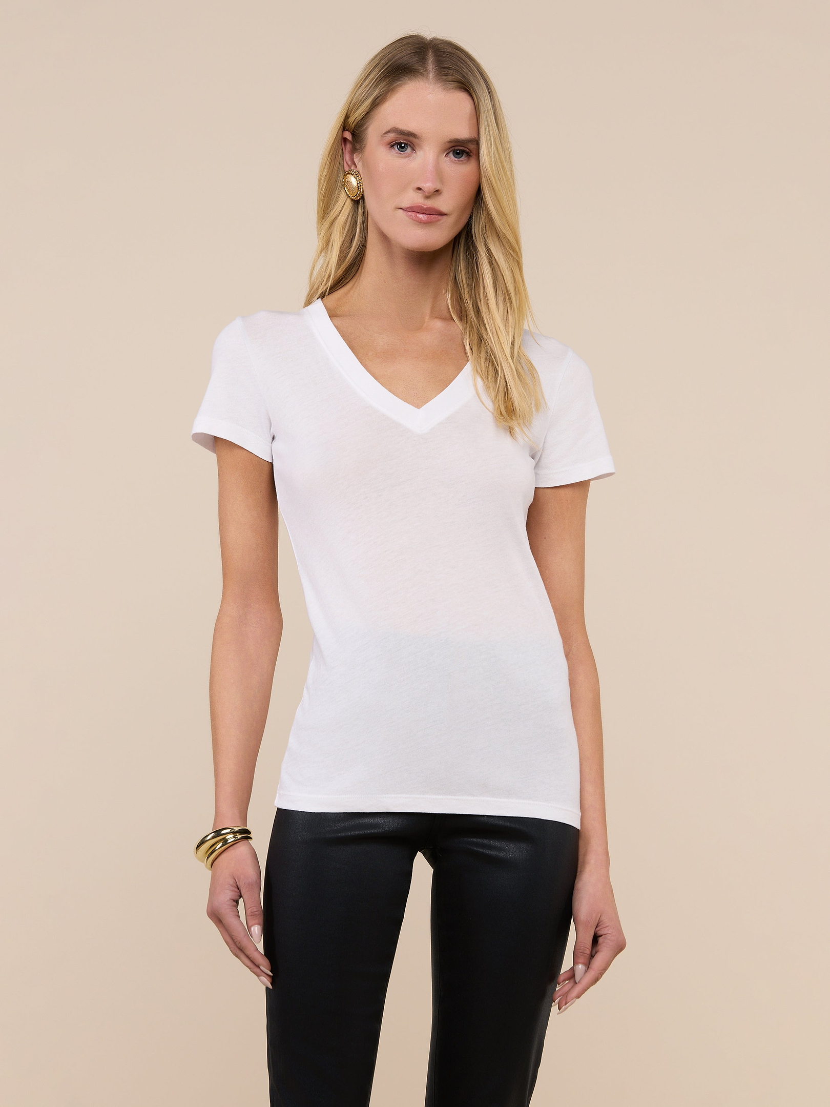 A woman poses against a plain background wearing the Becca Cotton V-Neck Tee, crafted from 100 cotton in white, paired with black pants, gold hoop earrings, and bracelets, her arms relaxed at her sides.