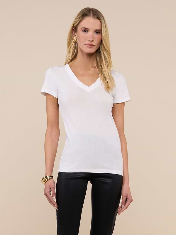 A woman poses against a plain background wearing the Becca Cotton V-Neck Tee, crafted from 100 cotton in white, paired with black pants, gold hoop earrings, and bracelets, her arms relaxed at her sides.