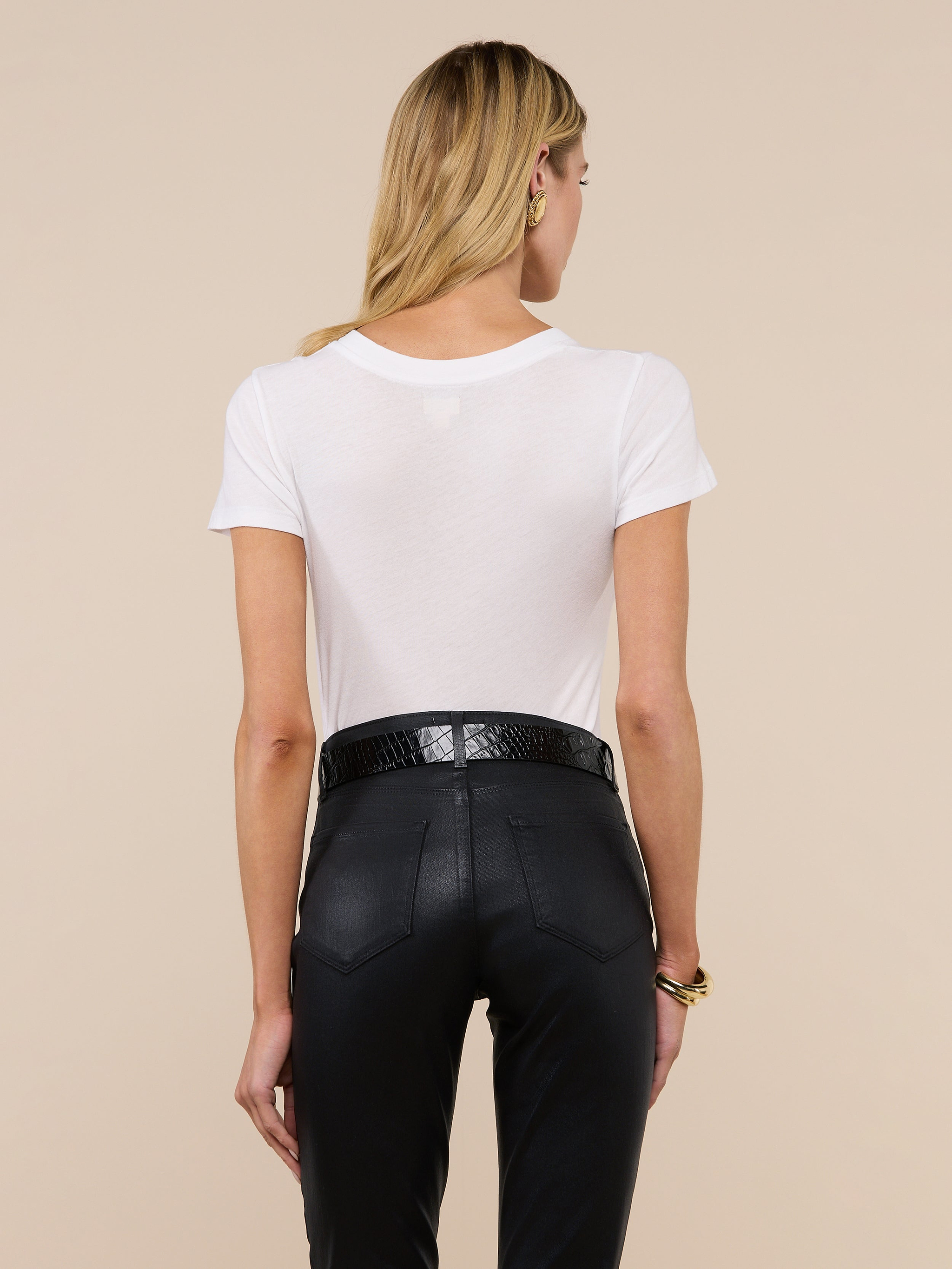 A woman with long blonde hair is seen from behind, wearing the Becca Cotton V-Neck Tee in white, tucked into high-waisted black pants with a black belt, against an off-white background.