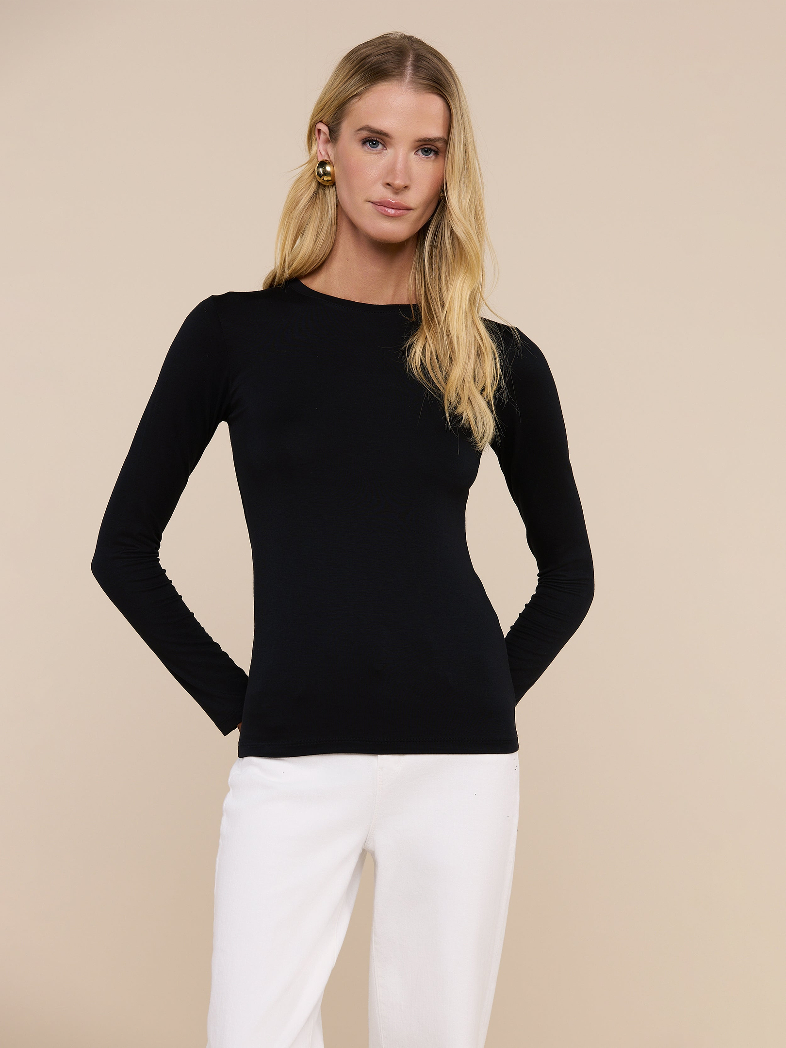 A woman with long blonde hair wears the fitted black Tess Long Sleeve Tee and white pants, standing against a plain light background with her hands behind her back.