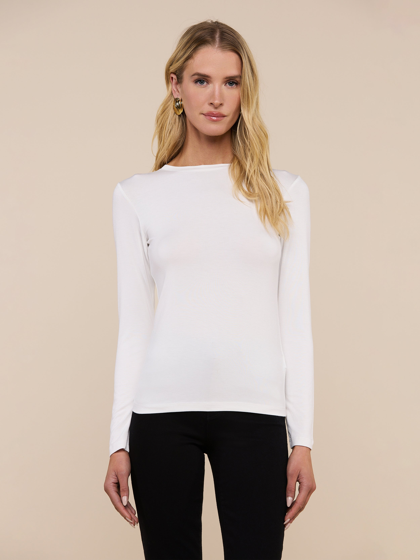 A woman with long blonde hair wears the Tess Long Sleeve Tee in white and black pants, standing facing forward against a plain, light background.