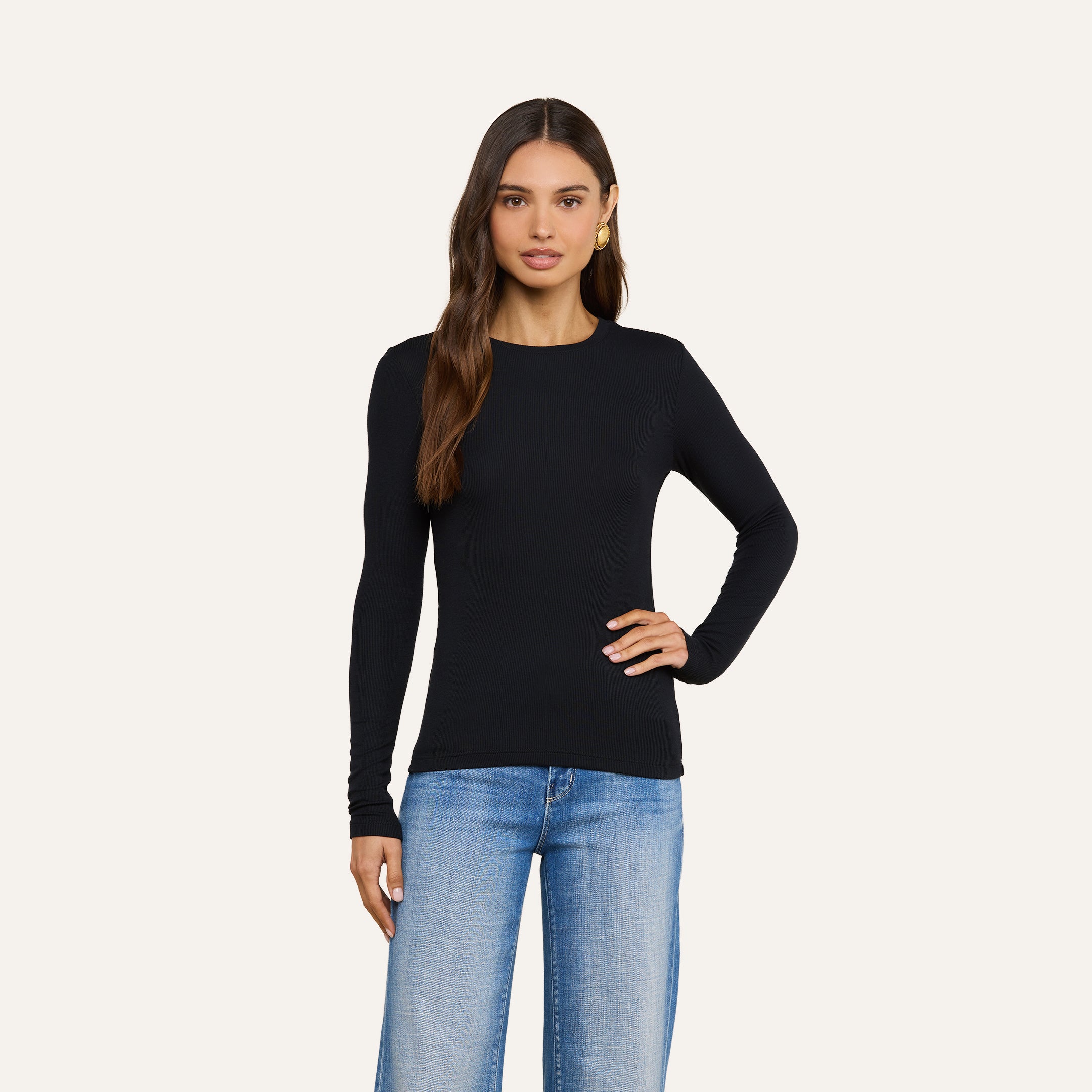 A woman with long brown hair wearing the Tess Long Sleeve Tee in black, paired with light blue jeans, stands with one hand on her hip against a plain light background.