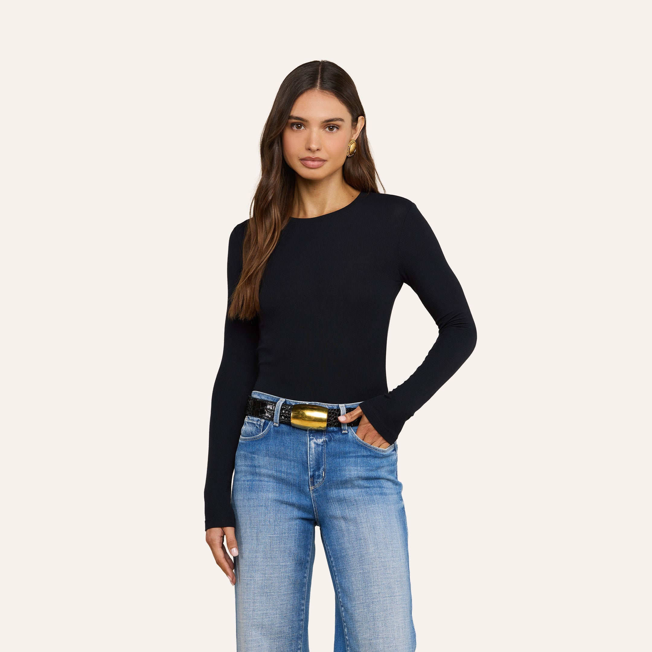 Woman with long brown hair wearing the Tess Long Sleeve Tee in black, styled with blue jeans, a black belt featuring a gold buckle, and gold hoop earrings, standing against a plain light background with one hand in her pocket.