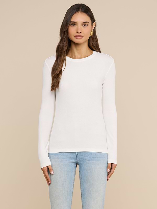 Wearing the Tess Long Sleeve Tee in white, a woman with long brown hair and a neutral expression stands facing forward against a light background, paired with light blue jeans.