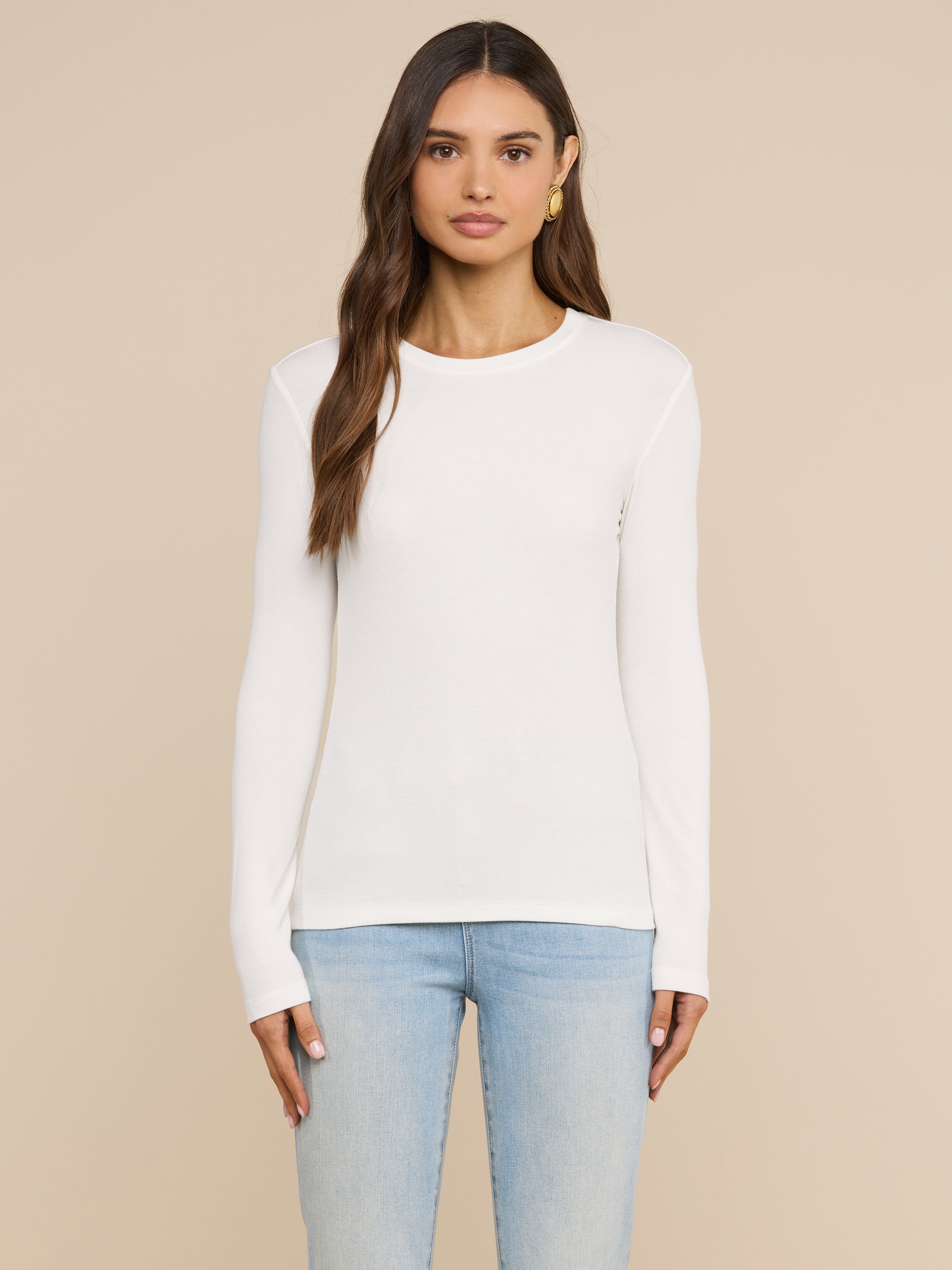 Wearing the Tess Long Sleeve Tee in white, a woman with long brown hair and a neutral expression stands facing forward against a light background, paired with light blue jeans.
