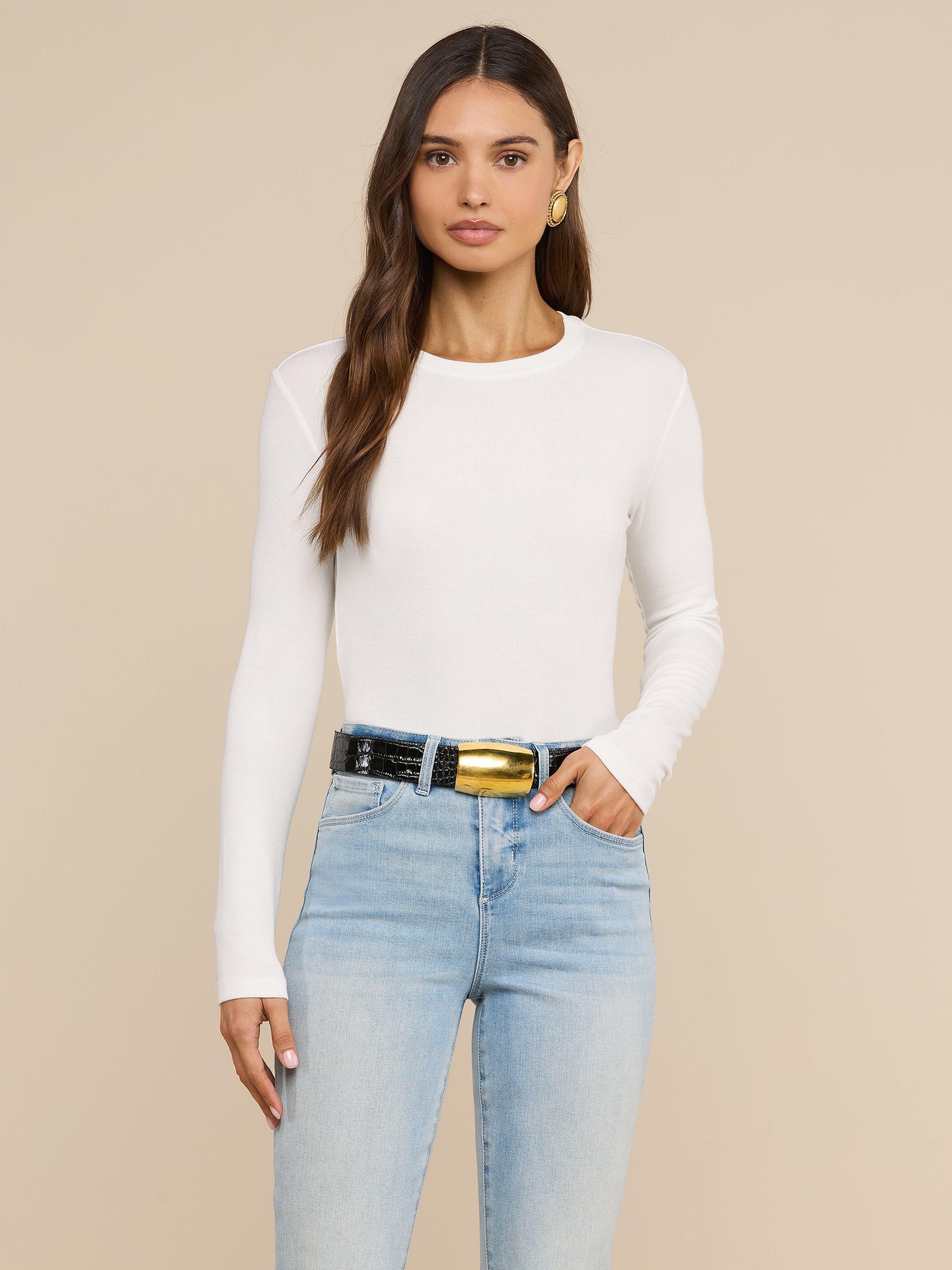 A woman with long brown hair stands against an off-white background wearing the Tess Long Sleeve Tee, a slim-fit white crew neck top, paired with light blue jeans, a black belt featuring a large gold buckle, and gold hoop earrings.
