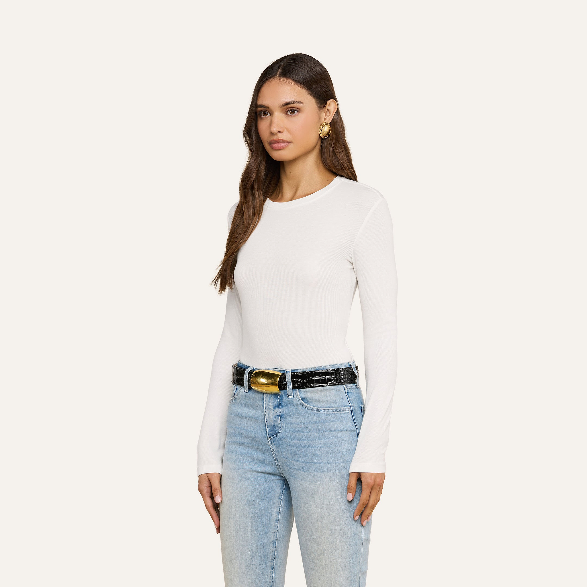 A woman with long brown hair wears the Tess Long Sleeve Tee—a slim-fit, ribbed micro modal white crew neck top—paired with light blue jeans, a black belt featuring a gold buckle, and large gold earrings. She stands against a plain light background, facing forward and slightly to the left.