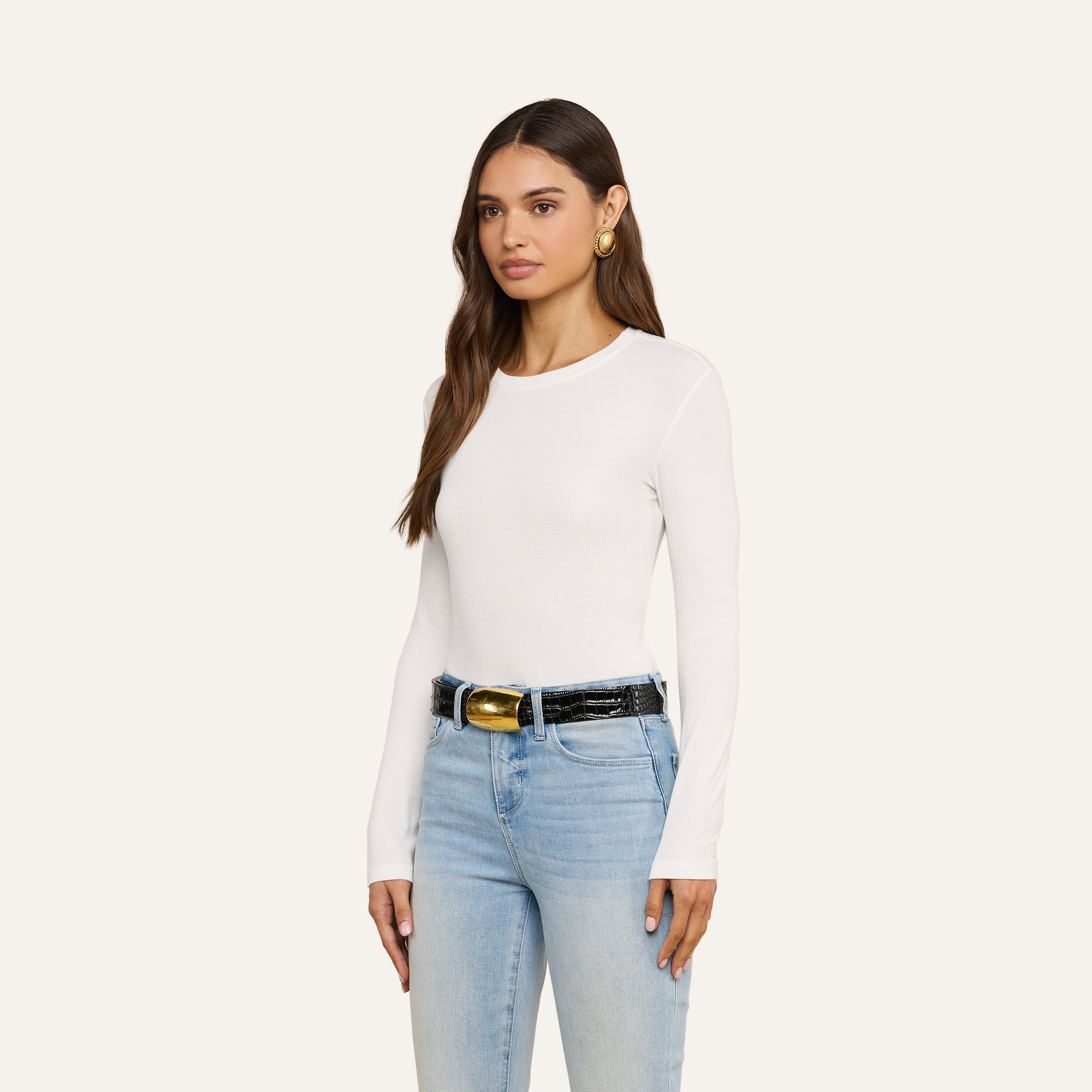 A woman with long brown hair wears the Tess Long Sleeve Tee—a slim-fit, ribbed micro modal white crew neck top—paired with light blue jeans, a black belt featuring a gold buckle, and large gold earrings. She stands against a plain light background, facing forward and slightly to the left.