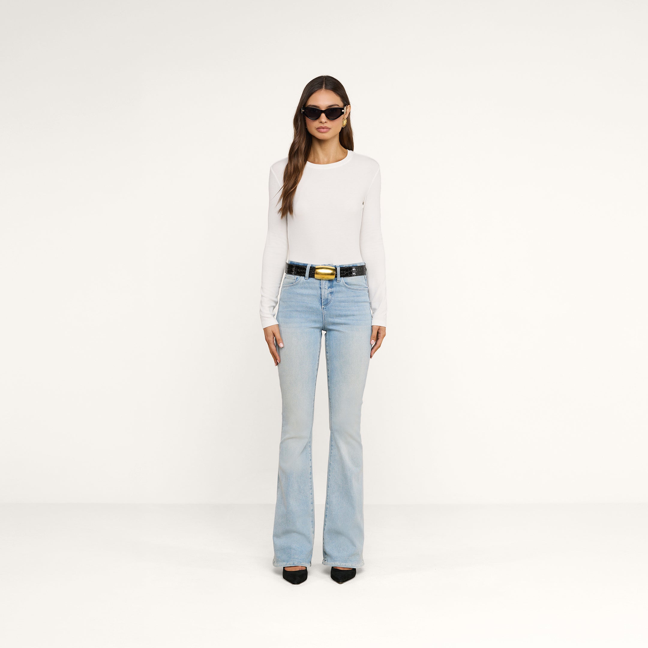 A woman wearing sunglasses, the Tess Long Sleeve Tee—a slim-fit white crew neck top made of ribbed micro modal—light blue flared jeans, a black belt, and black shoes stands against a plain white background.