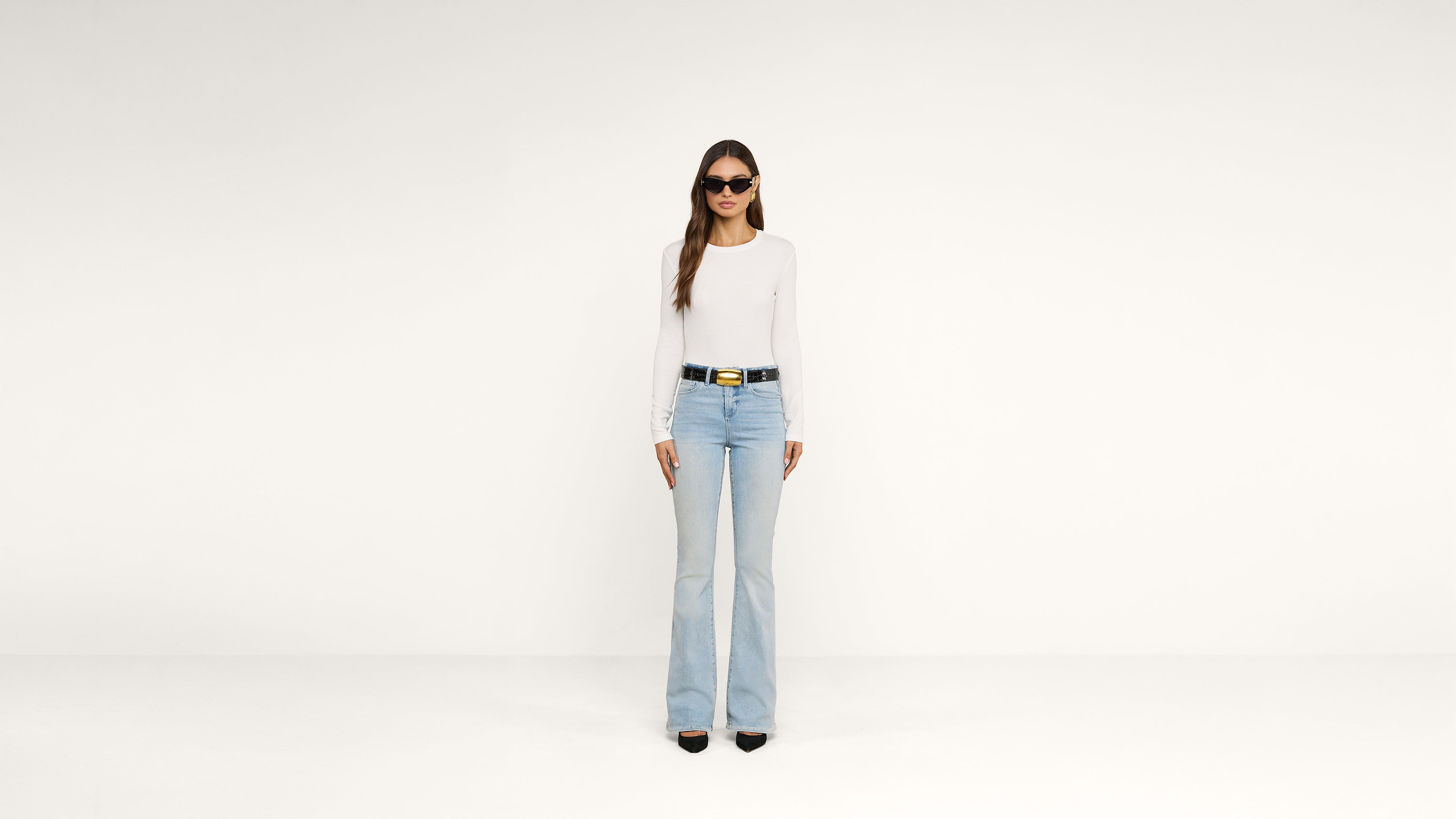 Woman in sunglasses, a white long-sleeve top, light blue flared jeans, and black heels stands against a plain white background.
