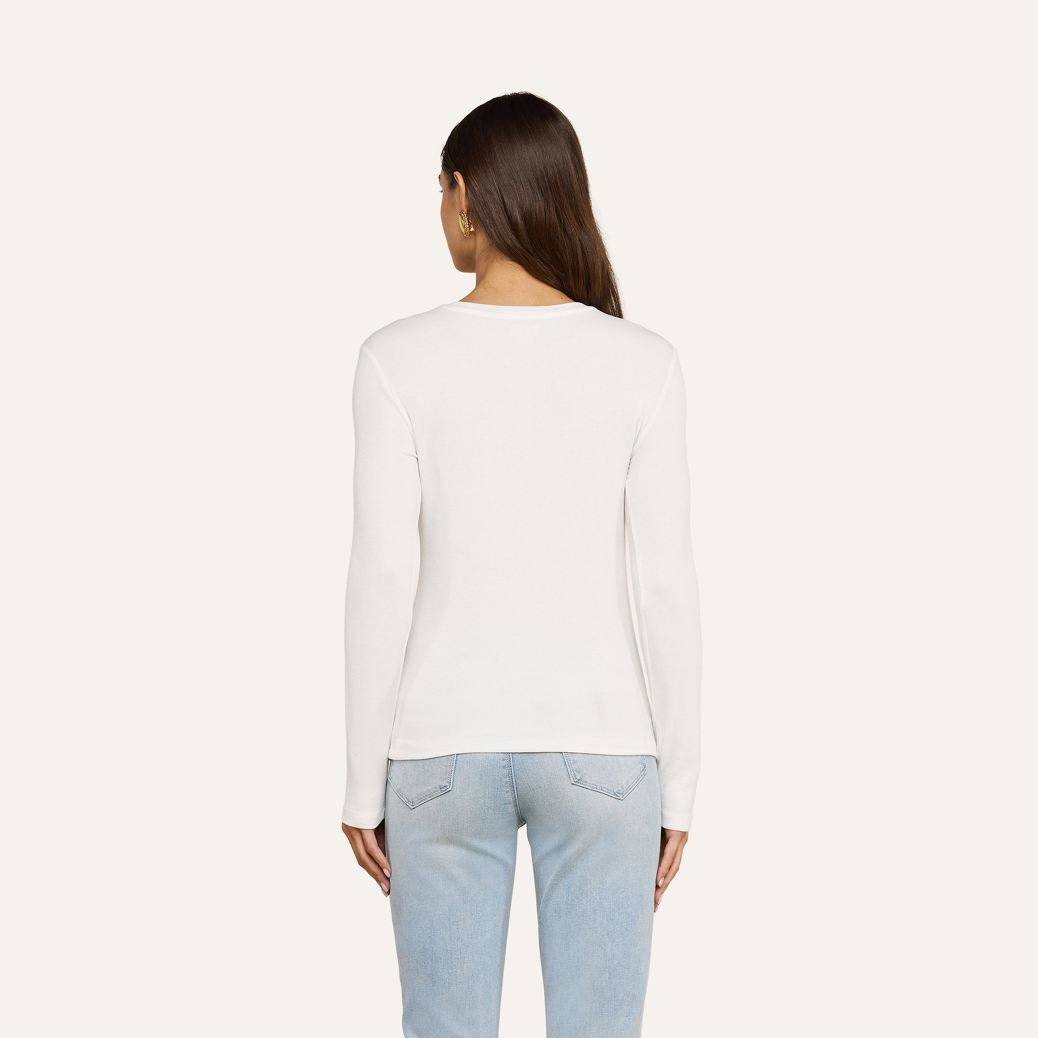 A woman with long brown hair is standing with her back to the camera, wearing the Tess Long Sleeve Tee in white and light blue jeans against a plain light background.
