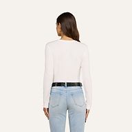 A woman with long brown hair is shown from the back, wearing the Tess Long Sleeve Tee in white, paired with light blue jeans and a black belt, standing against a plain light background.