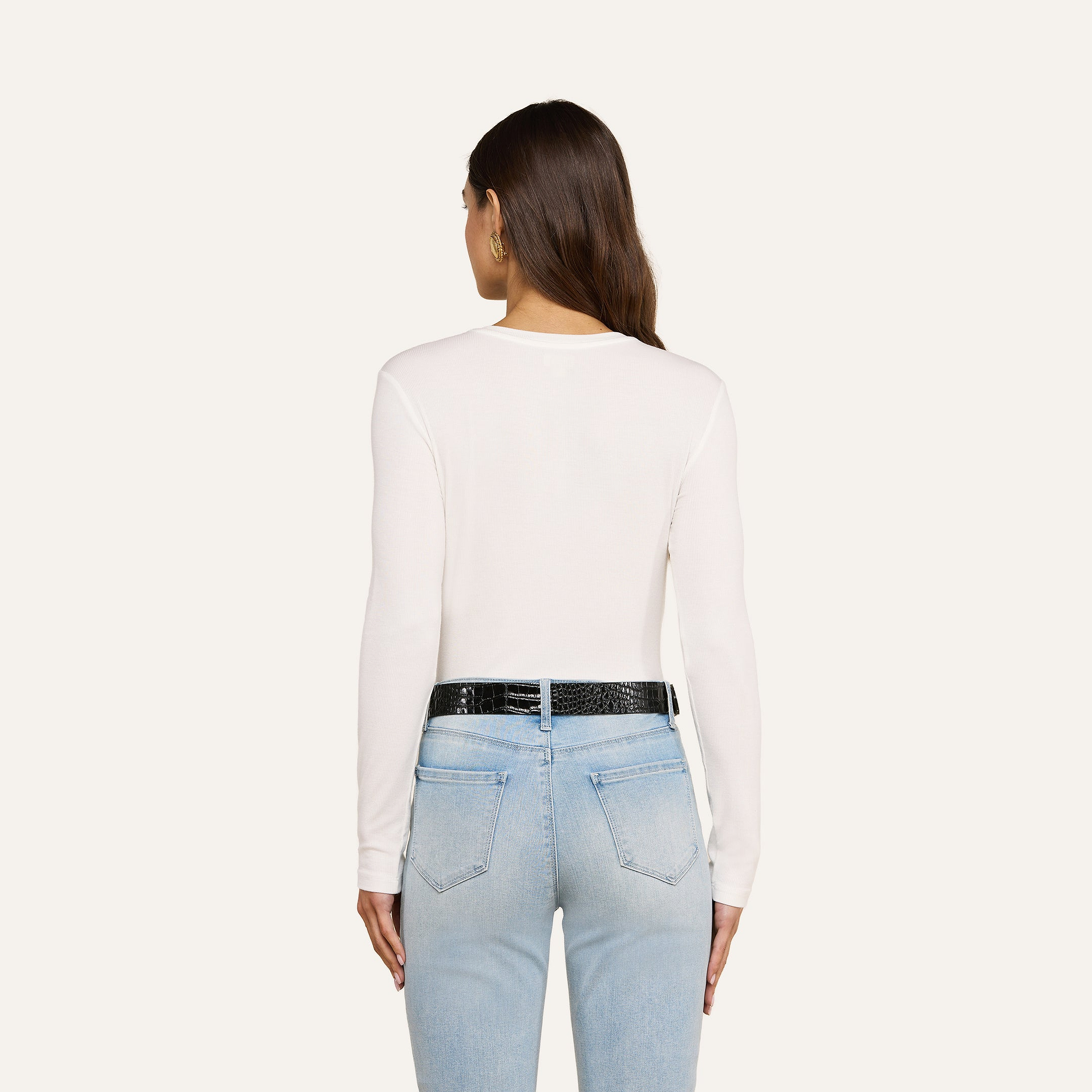 A woman with long brown hair is shown from the back, wearing the Tess Long Sleeve Tee in white, paired with light blue jeans and a black belt, standing against a plain light background.